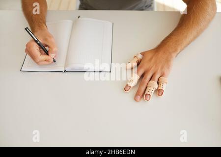 Man making notes in notebook wears plastic splints on broken or injured fingers of his hand. Stock Photo