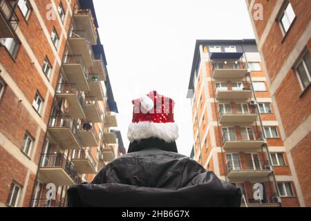 Back view of female person walking on the street with cmas hat on wudounded by block houses. Self isolation and quarantine. Holidays in a city lonely. Stock Photo