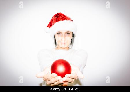 Woman in Christmas outfit holds in two hands red xmas decoration shiny ball gift. Happiness and sharing of love  and hope on celebrations Stock Photo