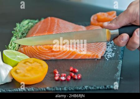Hand chef using knife slice raw salmon on chopping block. Salmon fillet steak and seasonings on black cutting board. Stock Photo