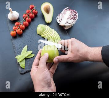 Hand with knife cutting out an apple core on a dark chopping board, fresh juicy green apple sliced scattered on the black background Stock Photo