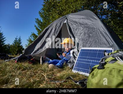 Adorable male child in construction helmet using mobile phone while sitting in tourist tent near portable solar panel. Cute kid with modern cellphone resting in camp tent outdoors. Stock Photo