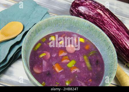 View on isolated purple rustic soup bowl with leek, zucchini and sausages, color comes from black carrots Stock Photo
