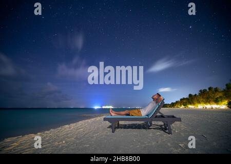 Man lying on sunbed on sand beach and looking up to stars on night sky. Stock Photo