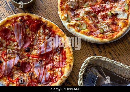 Top view of two fresh baked homemade sliced pizzas with cheese and ham topping served with kitchenware on wooden background. Stock Photo