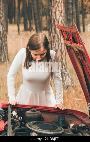 Chernigov, Ukraine - November 10, 2020: A girl in a white wedding dress looks at the car engine. Confused girl near the car Stock Photo