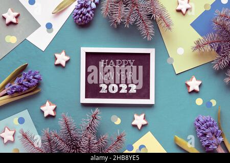 Happy New Year 2022 text on black board. Green layered geometric paper background with star cookies, blue hyacinth flowers and fir twigs. Winter