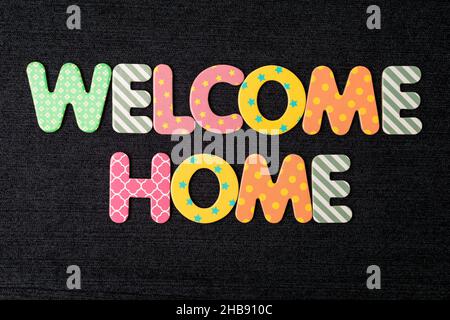 Card with Welcome Home words made from mixed vivid colored wooden letters on a textured dark black textile material that can be used as a message Stock Photo