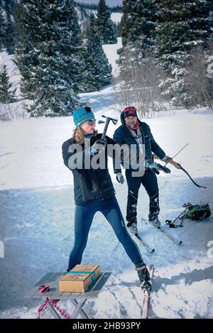 A slim woman stands in the snow on cross country skis, prepares to throw an ax, a man behind her holds an archery bow. Stock Photo