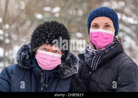 Elderly senior woman with her younger daughter in warm winter clothes, both of them wearing pink single use disposable virus face mask Stock Photo