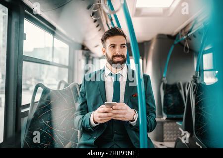 Smiling ambitious bearded businessman in turquoise suit sitting in public bus and using smart phone. If you want to fly, give up everything that weigh Stock Photo