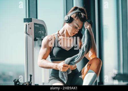 Serious muscular strong female bodybuilder with ponytail and headphones wiping sweat while sitting in gym next to window. The start is what stops most Stock Photo