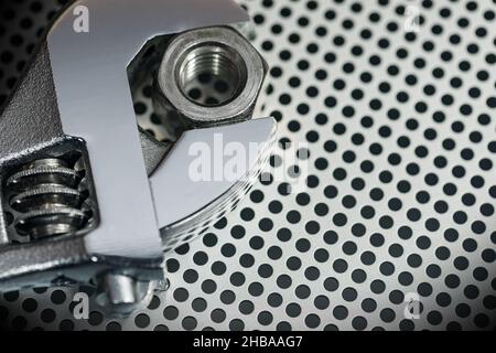 Extreme close-up of a modern stainless steel adjustable wrench and a bolt on a metallic background with many holes and copy space, photography. Stock Photo