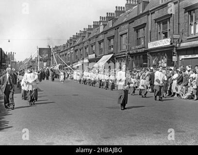 A Whit Walks procession in Union Street, Oldham, Greater Manchester, Lancashire, England, UK c.1960. The large banner has an image of a saint and the words ‘Pray for us’ on it. A man carries a cross ahead of choirboys. The street’s shops included a newsagent (advertising the Manchester Evening Chronicle) and a branch of the Failsworth Building Society. The Church of England religious event traditionally took place on Whit Friday, with children heavily involved along with brass and silver bands. This is taken from an old black and white negative – a vintage 1950s/60s photograph. Stock Photo