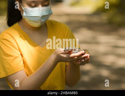 A teenage girl with a ball python in her hand, a girl in a mask and a yellow shirt stand in the garden. Stock Photo