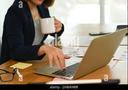 Woman in suit presses, hand on the laptop keyboard and holds coffee with the other hand, a young woman's hand is using laptop keyboard, drinking coffe Stock Photo