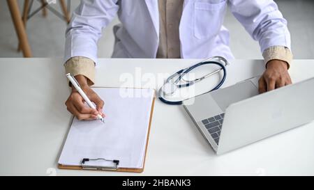 A medical professional in a lab coat holds a pencil on an empty file and operates a laptop, The stethoscope is placed on the table. Stock Photo