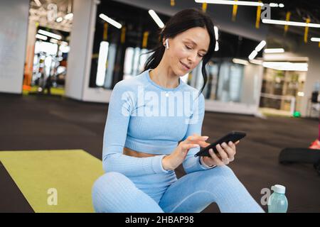 Joyful fitness woman in wireless headphones sitting on floor in gym and using smartphone Stock Photo