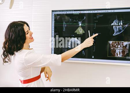 Beautiful senior woman dentist in white uniform and red belt smiling showing a snapshot of the teeth on the screen in her office in the dental clinic. Stock Photo