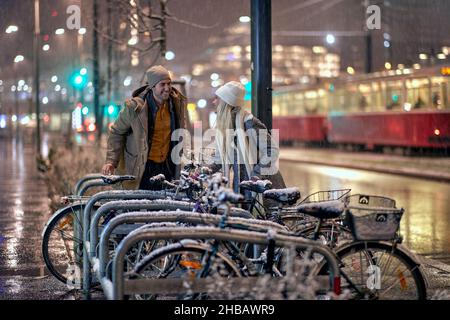 A young couple is preparing to ride bikes in a cheerful manner during a snowy night in the city. Christmas, New Year, holiday, love Stock Photo