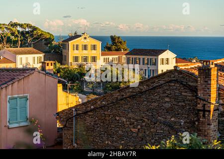 A cream-coloured villa with a curved gable and red shutters stands among pines above Bormes-les-Mimosas, Var department, France, in the evening light, against the open view of the Mediterranean Sea; a brick chimney rises above a dry-stone wall in the foreground. Stock Photo