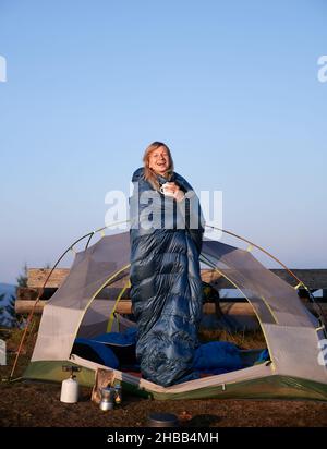 Sincere laughter of cheerful girl in sleeping bag who standing in tent and drinking coffee, on the background of cloudless light blue autumn sky.