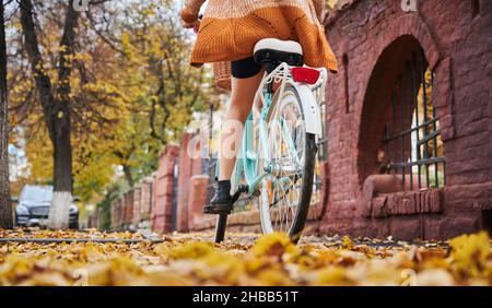 Rear view and close up young woman riding on blue bicycle in centre city in autumn park. Concept of riding on blue bicycle in middle fallen leaves in autumn. Stock Photo