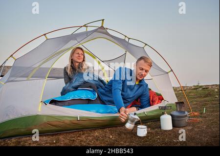 Charming young woman hiker sitting inside tourist tent and smiling while man pouring tea into cup. Happy couple travelers resting in comfortable camp tent in mountains. Concept of relationships. Stock Photo