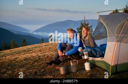 Man and woman resting at campsite in the mountains. Breakfast of two campers outdoors on background of beautiful mountain hills covered green forests. Concept of hiking, camping and relationships. Stock Photo