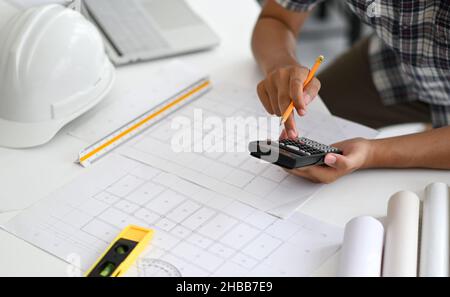 Architects are using a calculator to estimate the cost of house plans, house plans, safety helmet and office equipment placed on the desk. Stock Photo