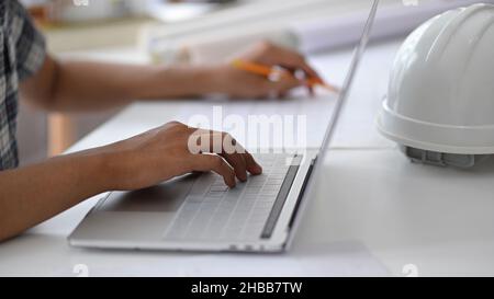 Young man hand is using a laptop keyboard, a safety helmet on the desk, Architect designing a house. Stock Photo