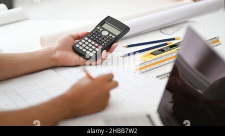Hand holding a calculator and pencil checking house plans, engineers are calculating the house scale, house plans and laptop on the desk. Stock Photo