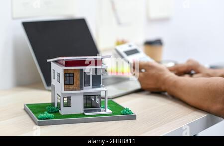 Model house on a table with blurry a person background using calculator and a laptop on the table, real estate expenses. Stock Photo
