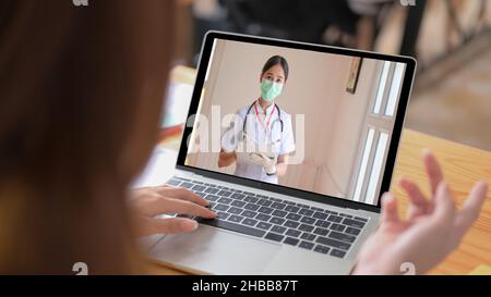 Sick woman uses a laptop video call to get advice from a medical professional, holding a tablet and wearing a mask. Stock Photo