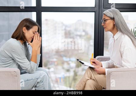 Worried young woman patient sitting at a psychologist's therapist appointment and telling about mental problems, attentive mature female doctor is listening and making notes. Psychotherapy concept Stock Photo