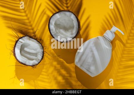 Minimalistic beauty still life. Two halves of chopped coconut and white bottle of cream with shadows from palm leaves on yellow background. Creative f Stock Photo