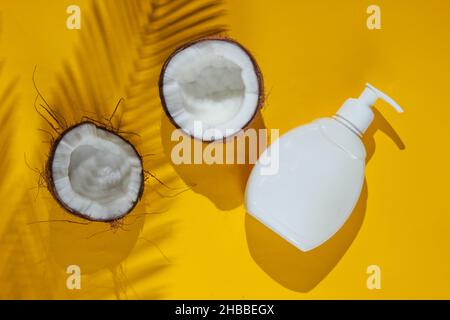 Minimalistic beauty still life. Two halves of chopped coconut and white bottle of cream with shadows from palm leaves on yellow background. Creative f Stock Photo