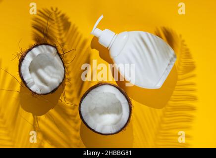 Minimalistic beauty still life. Two halves of chopped coconut and white bottle of cream with shadows from palm leaves on yellow background. Creative f Stock Photo