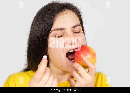 A woman bites a red apple while holding a multivitamin pill in her hand Stock Photo