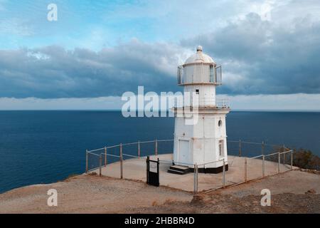 The white lighthouse stands on the mountain against the background of the sea and the stormy sky with dark clouds. Meganom lighthouse in Crimea. Stock Photo