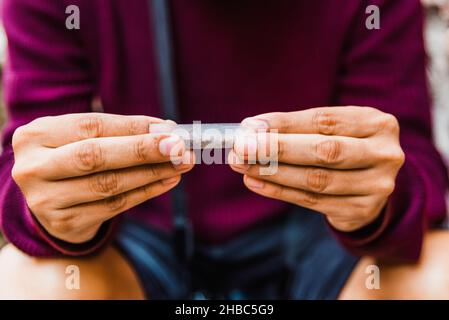 A young man uses cigarette paper to roll a cigarette on a street. Stock Photo