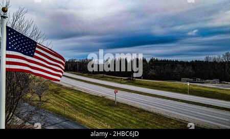 US Flag flows in the wind with a moody sky above during autumn. Country highway in the background with trees. November 2021