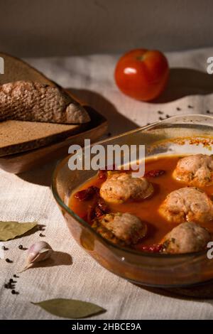 prepared meatballs in sweet tomato sauce in a glass baking dish, next to rye bread, linen tablecloth, tomato. Low key Stock Photo