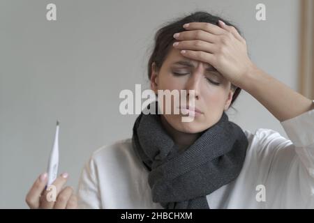 Unhealthy italian woman in warm scarf with eyes closed, holds digital thermometer Stock Photo