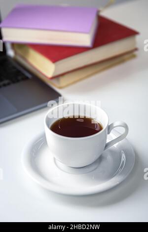 Cup of tea with saucer on the table near books and laptop. Stock Photo