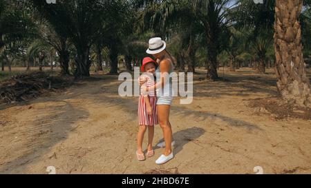 Woman tourist with plait walks looking around at growing young trees with lush leaves at oil palm farm elaeis guineensis on sunny day. Mom and daughte Stock Photo