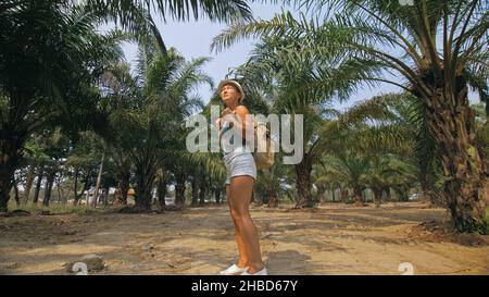 Woman tourist with plait walks looking around at growing young trees with lush leaves at oil palm farm elaeis guineensis on sunny day. Concept of exot Stock Photo