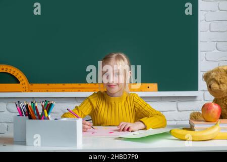 Back to school. Cute pupil girl drawing at the desk. Child in the class room with blackboard on background. Education, learning and children concept. Stock Photo