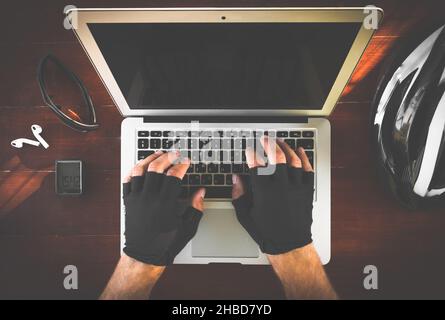 Two hands with gloves are typing on the keyboard of portable laptop with cycling accessories (helmet; cycle computer, earphones, glasses) layed on the Stock Photo