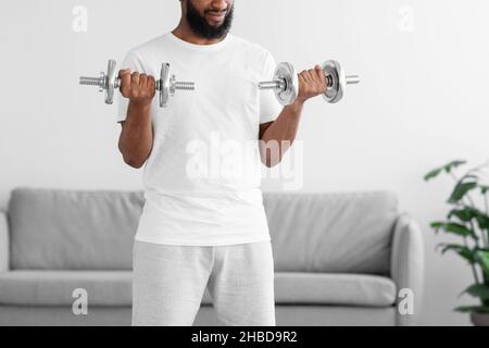 Unrecognizable millennial black athlete man in white sports clothes do exercise with dumbbells in living room Stock Photo